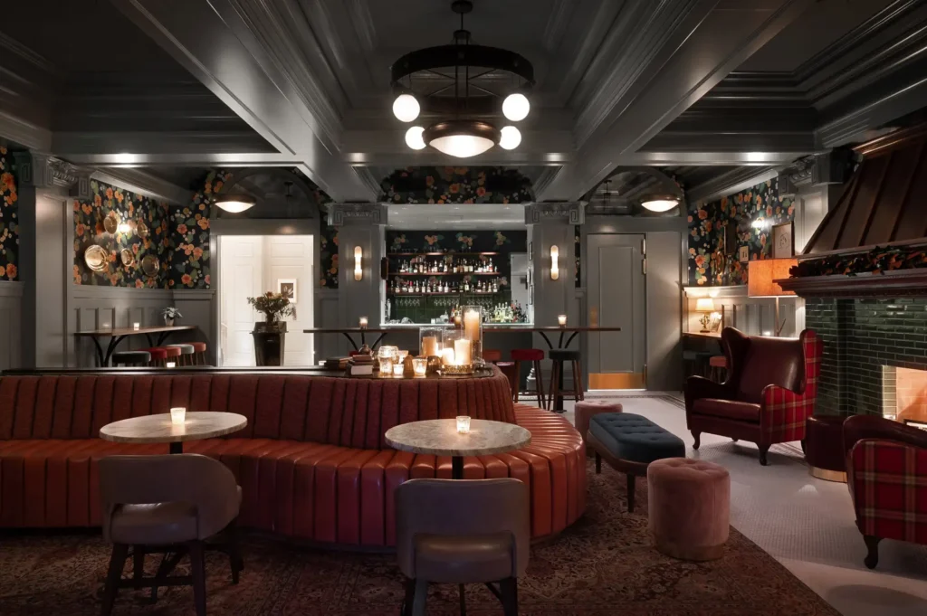 A cozy, dimly lit lounge with leather seating, marble tables, a bar stocked with bottles, floral wallpaper, chairs by a fireplace, pendant lights expertly installed by a Vancouver electrician, and candles on tables.