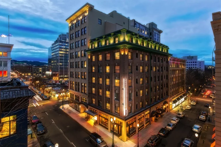 Nighttime city scene featuring a multi-story, well-lit building on a busy intersection with cars and lighted windows. An electrician in Vancouver, WA has masterfully ensured the surrounding buildings shine brightly against the darkening sky.