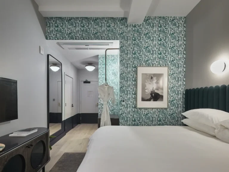 Modern hotel room with green patterned wallpaper, a bed with white linens, a wall-mounted TV installed by an electrician in Vancouver, WA, a full-length mirror, and a robe hanging near the entrance. A black-and-white framed photo adorns the wall.