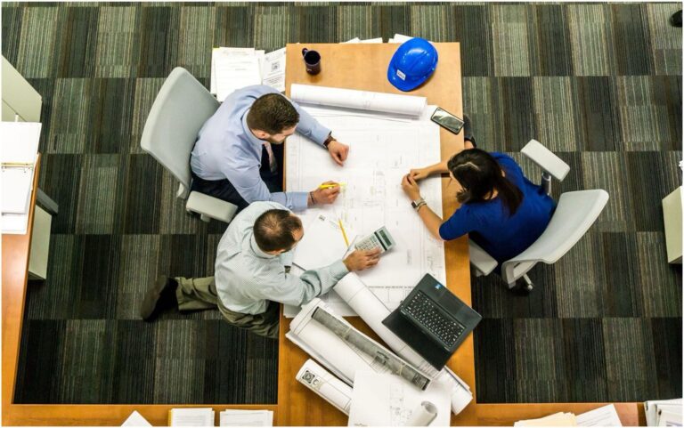 Three people working at a large desk with architectural plans, a laptop, and a blue hard hat. They are discussing electrical details and marking the plans with pens.