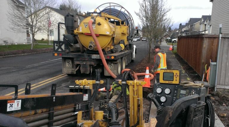 A construction crew, including a Vancouver electrician, working with a yellow vacuum truck and drilling equipment on a residential street. One worker in a high-visibility vest adjusts equipment near safety cones.