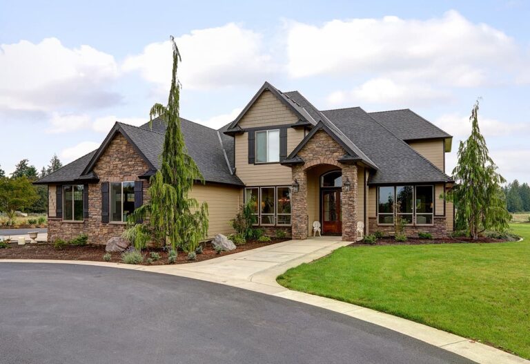 A large suburban house with a combination of stone and beige siding, multiple gables, a black shingle roof, and a well-maintained lawn, featuring trees at the front and a curved driveway. Whether you're looking for an electrician in Vancouver WA or just admiring the architecture, this home has it all.