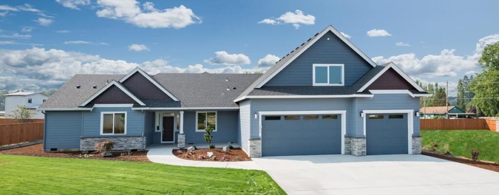 A blue, single-story house with a gabled roof, three-car garage, and a well-manicured lawn, set against a sky with scattered clouds. An electrician in Vancouver WA recently upgraded the home's electrical system to ensure it meets the highest safety standards.