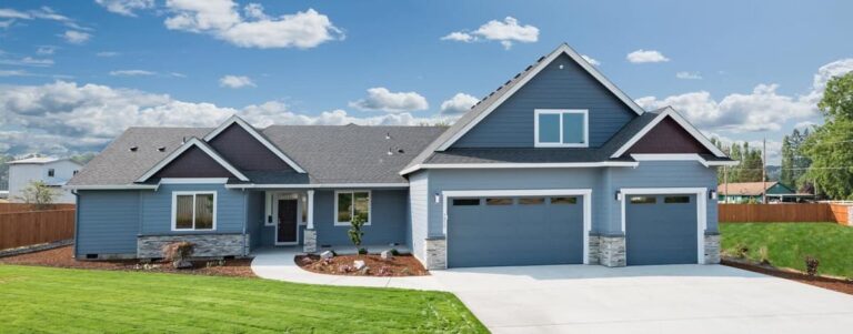 A blue, single-story house with a gabled roof, three-car garage, and a well-manicured lawn, set against a sky with scattered clouds. An electrician in Vancouver WA recently upgraded the home's electrical system to ensure it meets the highest safety standards.