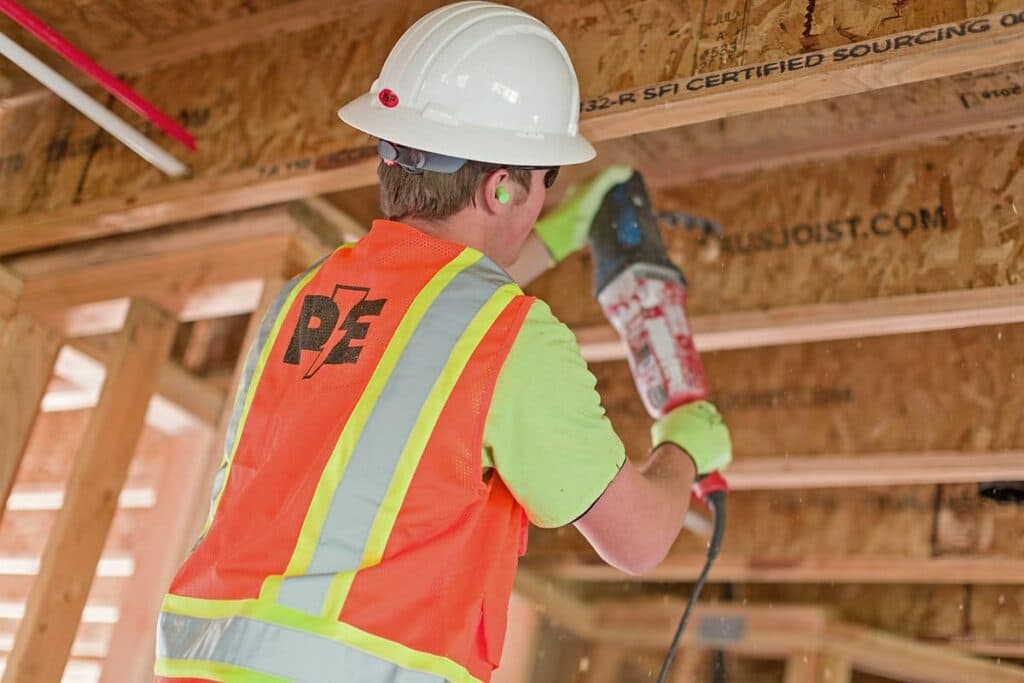A local electrician in Vancouver, WA, donning a hard hat, drills a hole in a wood beam.