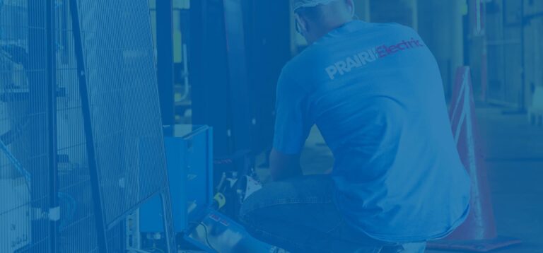 A person wearing a blue "PRAIRIElectric" shirt and a white safety helmet kneels while working on electrical equipment inside a facility, showcasing the dedication of a Vancouver electrician.