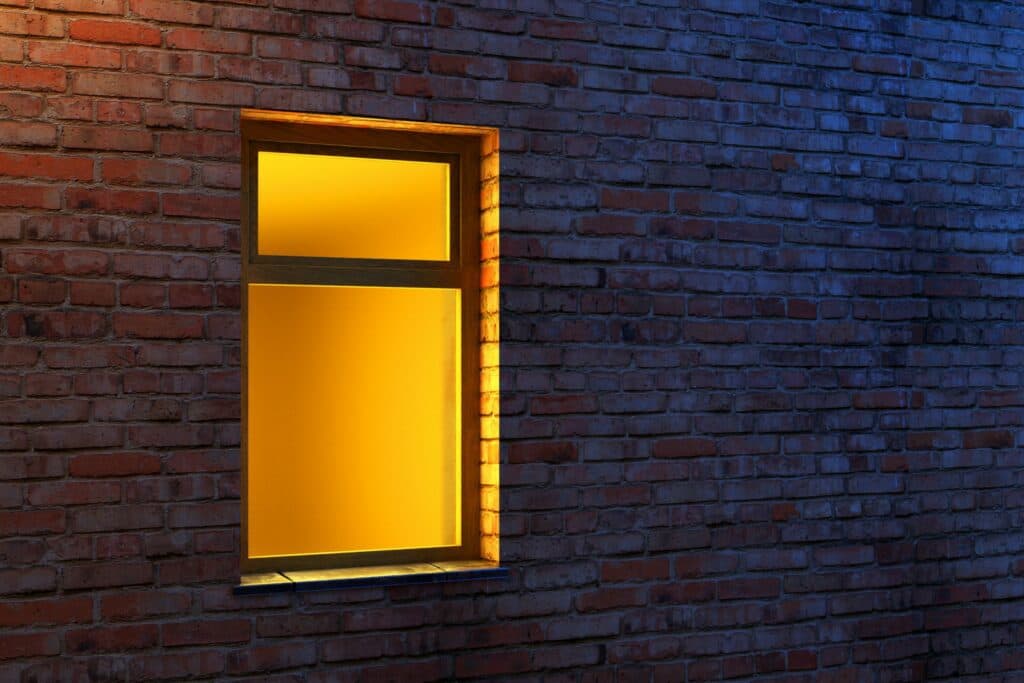 An illuminated window with yellow light in a brick building to illustrate How to Read an Electric Meter and Calculate Energy Usage.