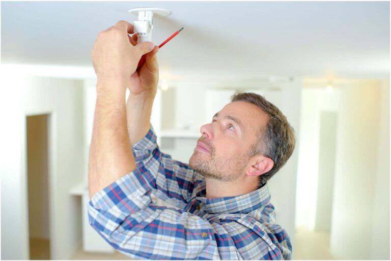 A man in a plaid shirt, resembling a Vancouver electrician, stands on a ladder, installing or repairing a ceiling light fixture while holding a red pencil.
