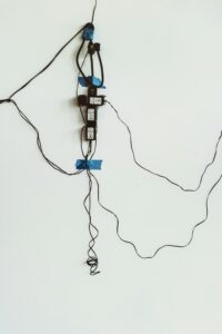 Several black electrical cords are taped to a white wall, some plugged into two visible electrical outlets, with blue tape securing them at multiple points. This setup might raise concerns during an electrical home inspection.