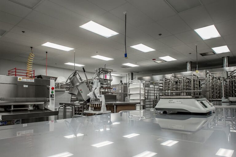 A large, well-lit commercial kitchen with stainless steel equipment, including ovens, racks, and workstations. Bright overhead lights installed by an electrician reflect on the shiny surfaces.