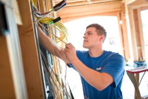 An electrician in a blue T-shirt is working on electrical wiring in a wooden structure, focusing intently on the task.