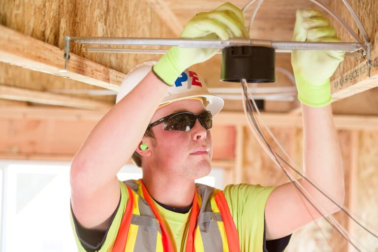 A construction worker wearing a hard hat, safety vest, gloves, and sunglasses is installing electrical wiring in a wooden framework. This skilled electrician in Vancouver WA ensures all installations meet safety standards.