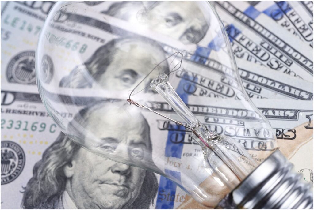 Close-up of a light bulb lying on a spread of U.S. one hundred dollar bills. The bulb is transparent, showing filament details, with Benjamin Franklin's portrait visible through the glass—an image that might intrigue any Vancouver electrician with an eye for innovation and craftsmanship.