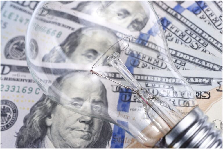 Close-up of a light bulb lying on a spread of U.S. one hundred dollar bills. The bulb is transparent, showing filament details, with Benjamin Franklin's portrait visible through the glass—an image that might intrigue any Vancouver electrician with an eye for innovation and craftsmanship.