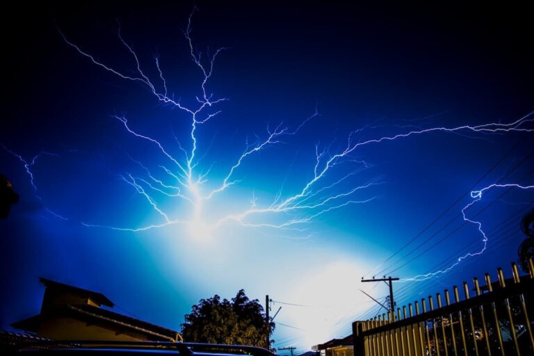 A night sky illuminated by multiple lightning bolts, with trees and rooftops silhouetted in the foreground, likely causing power surges, and reminiscent of the power harnessed by an electrician in Vancouver, WA.