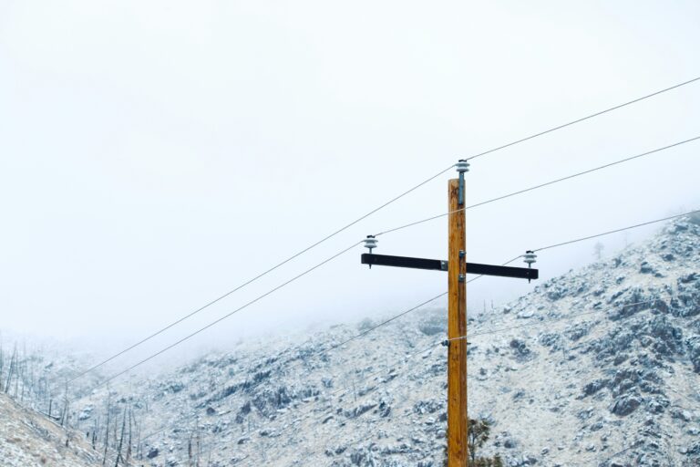 A wooden utility pole with power lines stands in front of a rocky, snow-dusted landscape under a cloudy sky.
