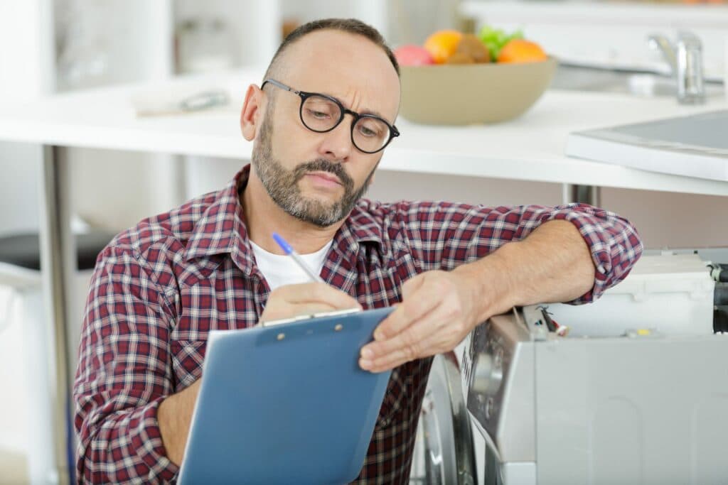 A local electrician from Vancouver, WA is inspecting a washing machine while maintaining records on a clipboard.